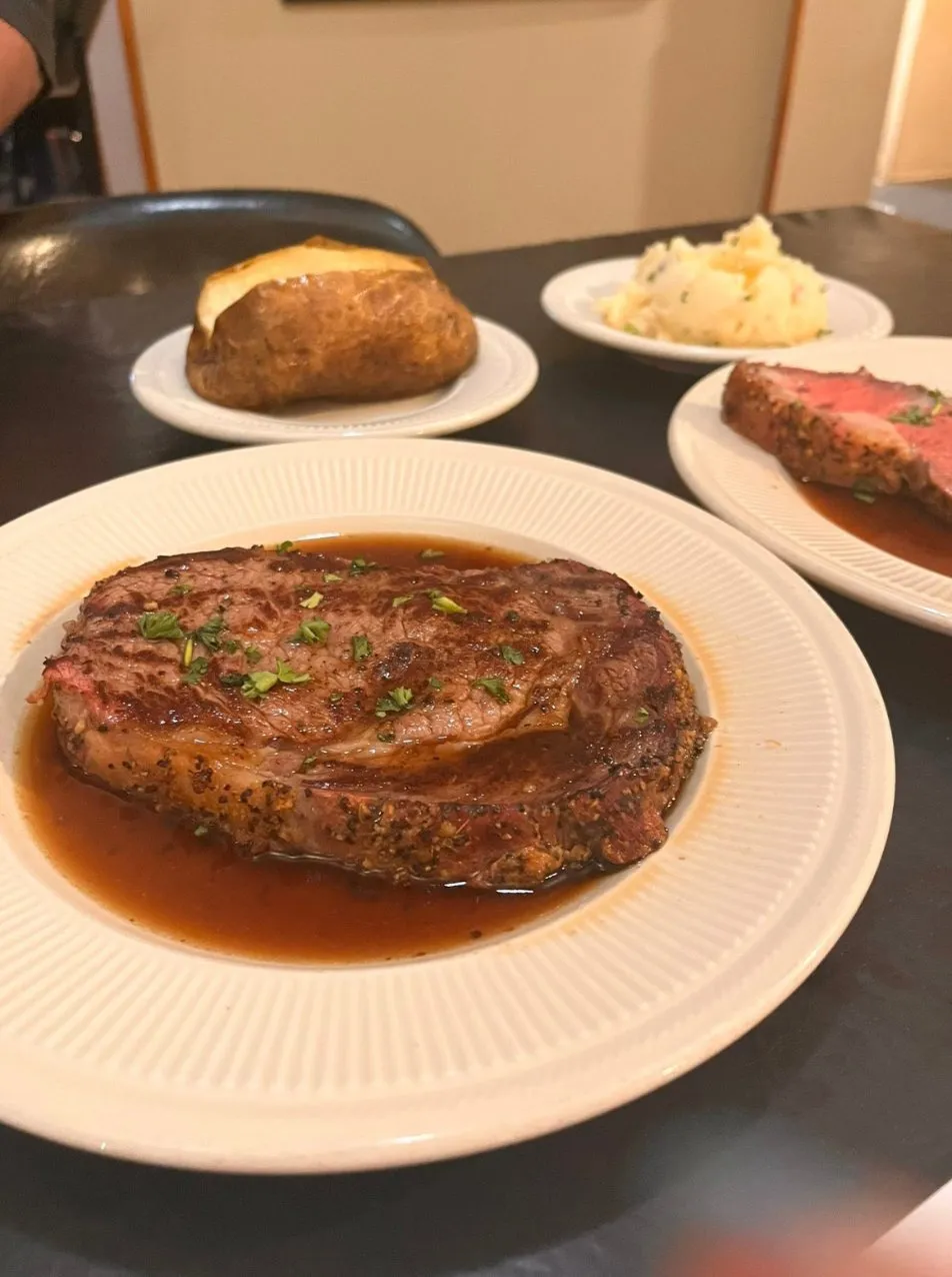 Peppercorn-crusted ribeye in au jus with a baked potato and prime rib behind it